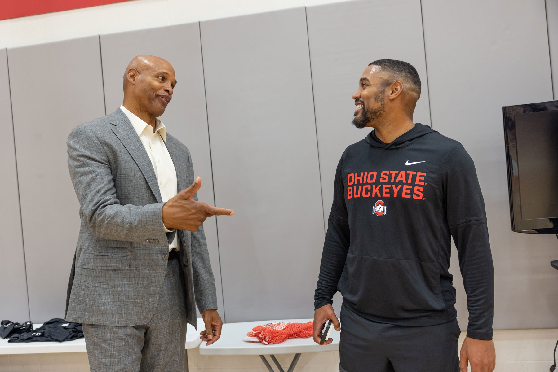 Kellogg and his adult son laugh as they talk in an indoor gym. Kellogg wears a suit and gestures with one hand. His son wears an Ohio State Buckeyes sweatshirt. 