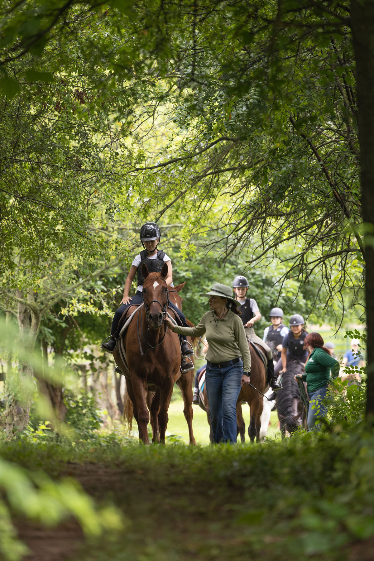 A group of people ride horses along a wooded trail, with one rider being guided by Lloyd walking beside the horse. Several other riders follow behind under the tree canopy.