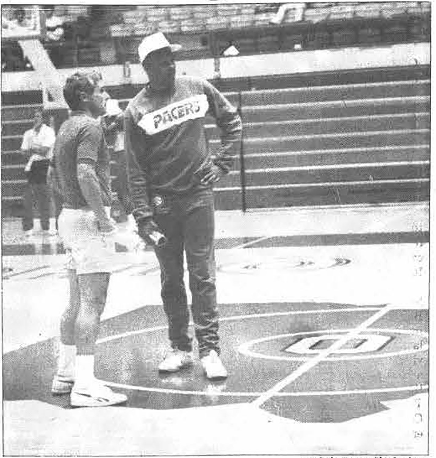 Kellogg wears a Pacers sweatshirt and baseball cap as he stands on a basketball court with Ohio State coach Gary Williams in St. John Arena. They appear to be watching college players outside of the photo. The photo comes from an old newspaper and is pixelated.