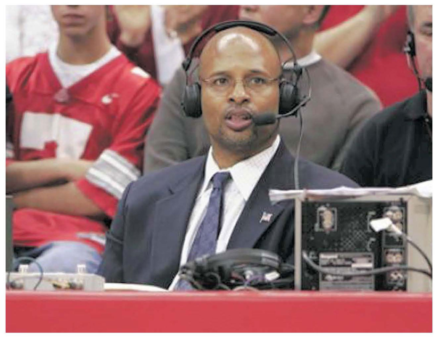 Kellogg wears a suit, tie, large headphones and a focused expression as he sits at a broadcast table courtside. Audio equipment and cables are positioned in front of the person, with spectators seated in the background.