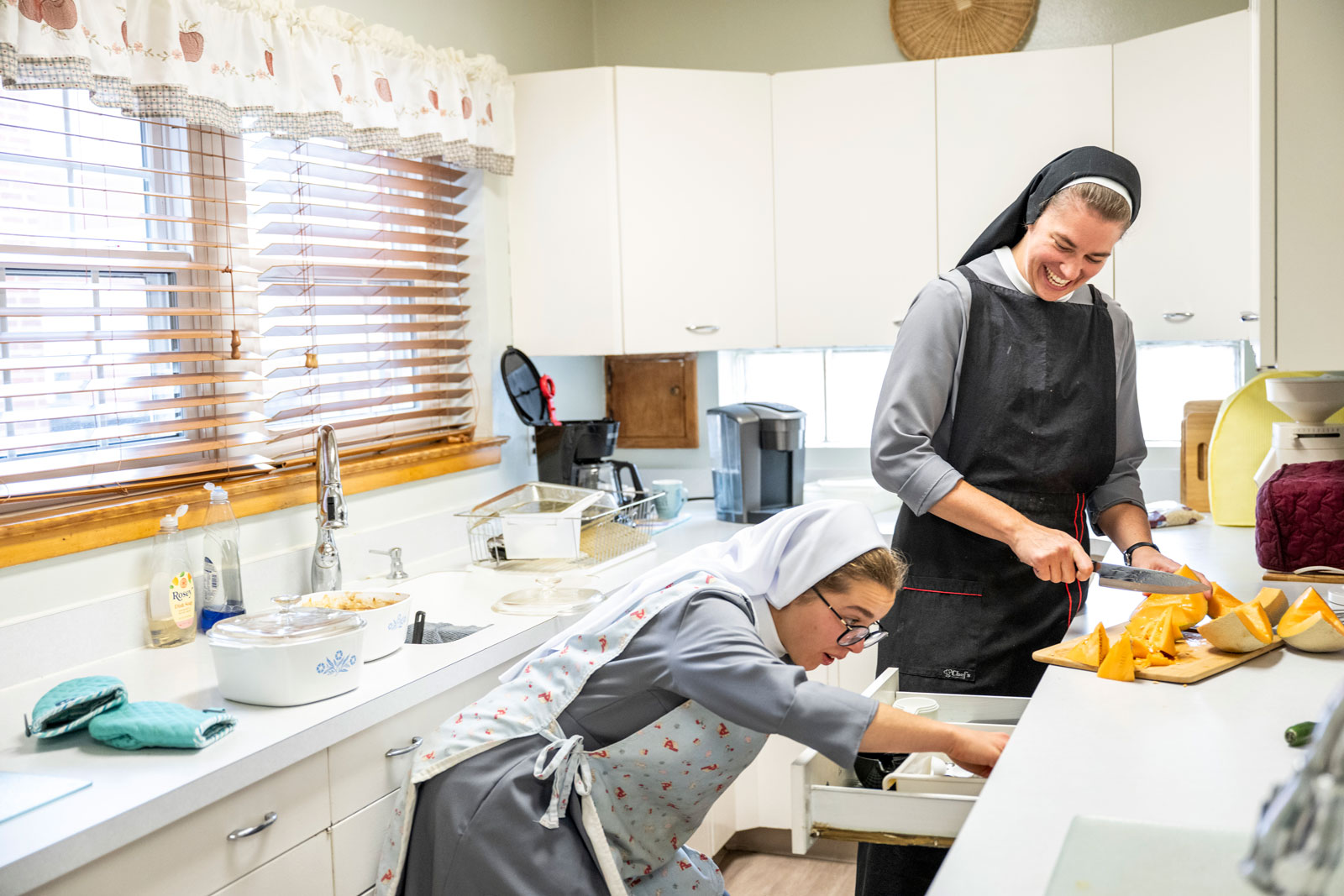 Two sisters cut up melons in a kitchen. Sister Xavier laughs as the other sister looks for something in a drawer.