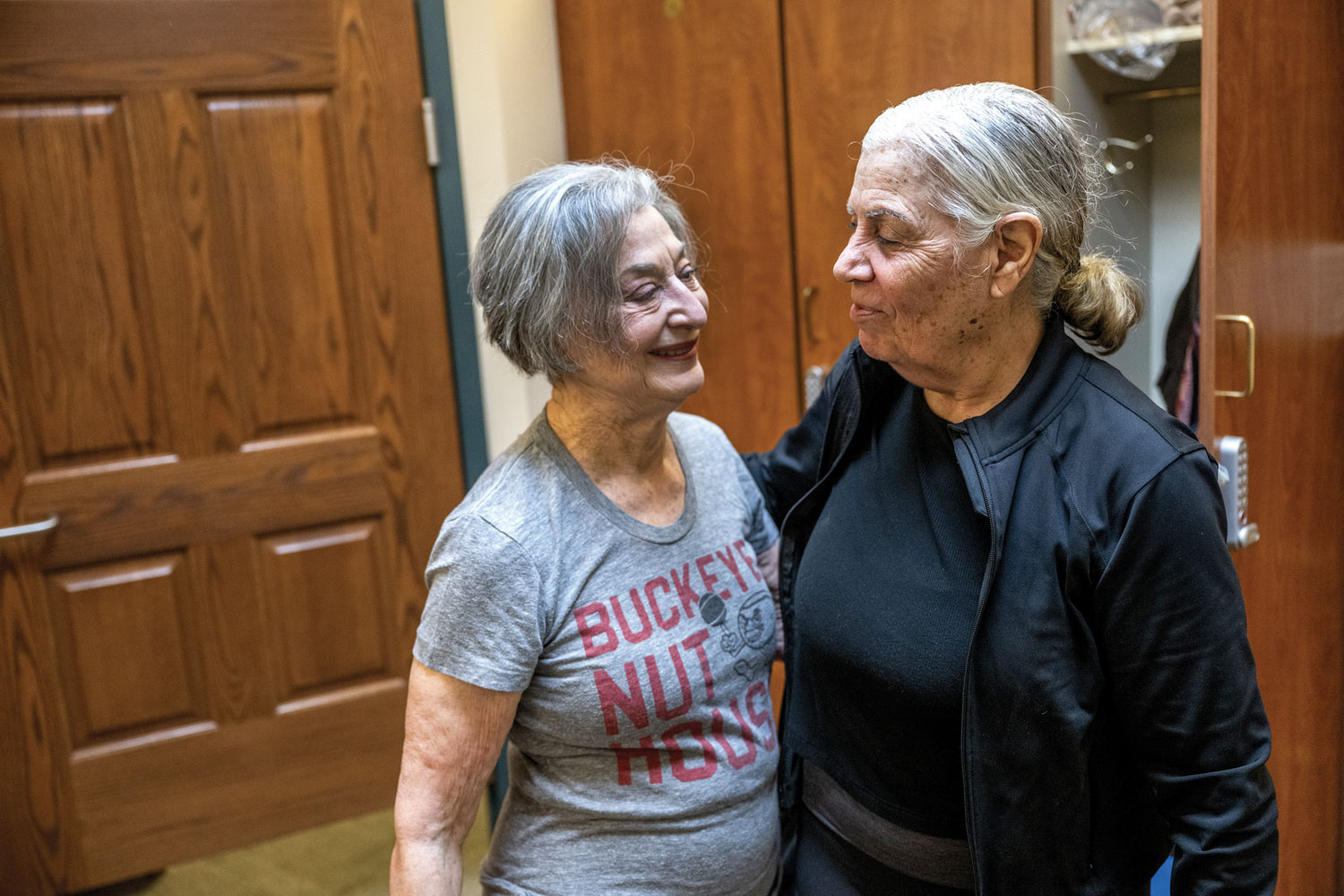 Flo Shleiferman Gurwin and a friend each wrap an arm around the other’s waist as they stand close close, chatting in a locker room with wooden cabinets. Flo is a white woman with gray hair just shorter than he chin and she wears a T-shirt with the words “Buckeye Nut House.” 
