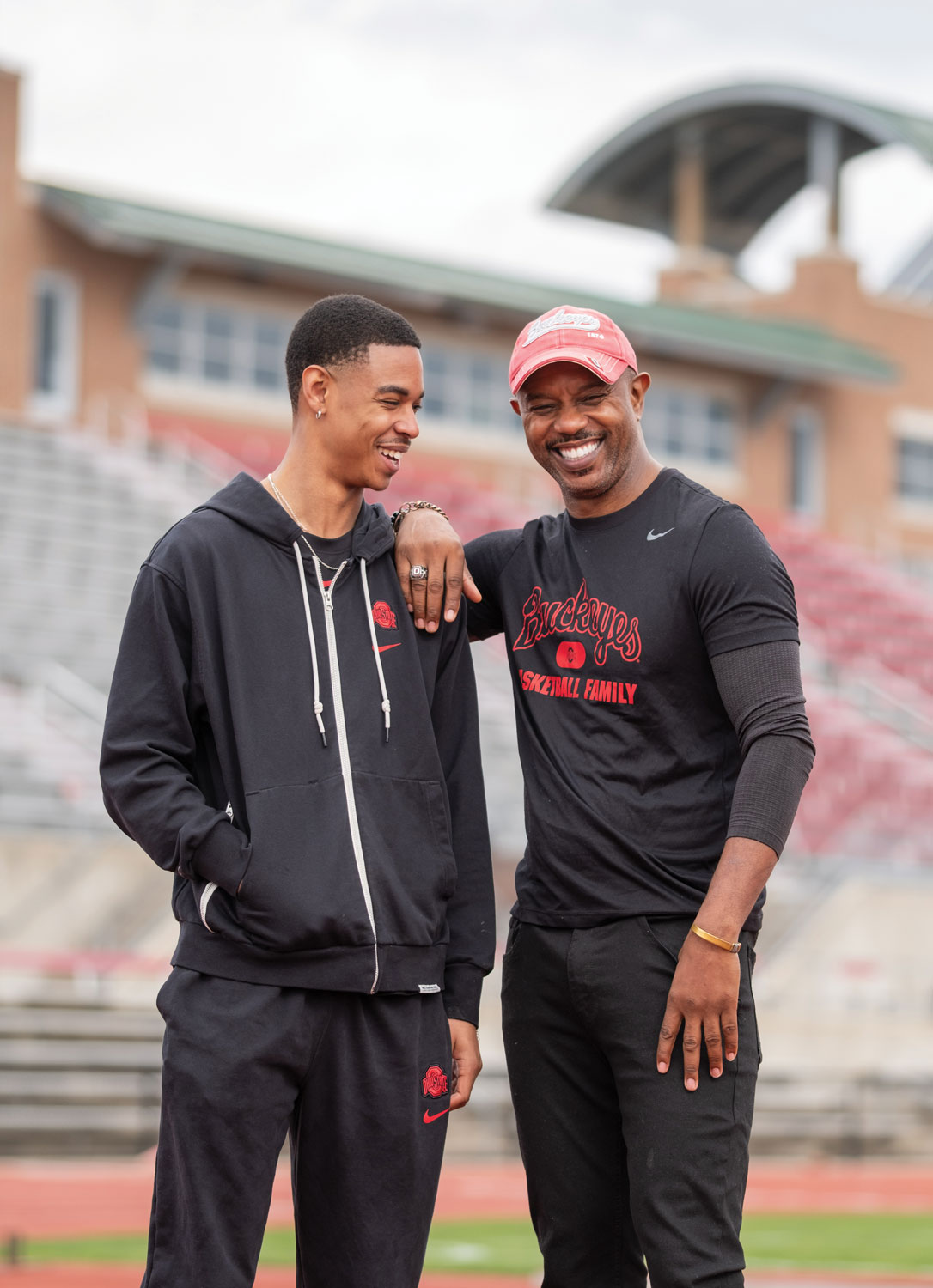 College-age son and father stand close together inside Jesse Owens Memorial Stadium, facing each other. Both are black men, laughing and wearing relaxed clothes. The father leans an arm on his son’s shoulder. Bleachers and a building appear in the background.