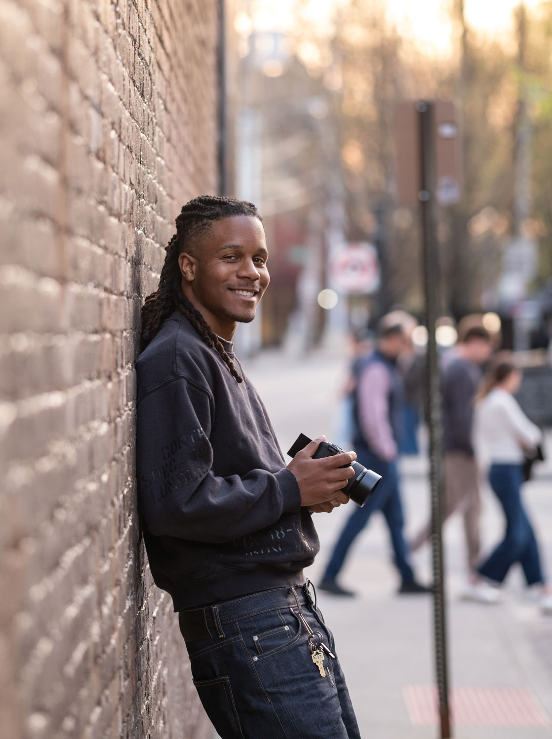 Justen leans back against a brick wall and grins while holding his video camera in this portrait.