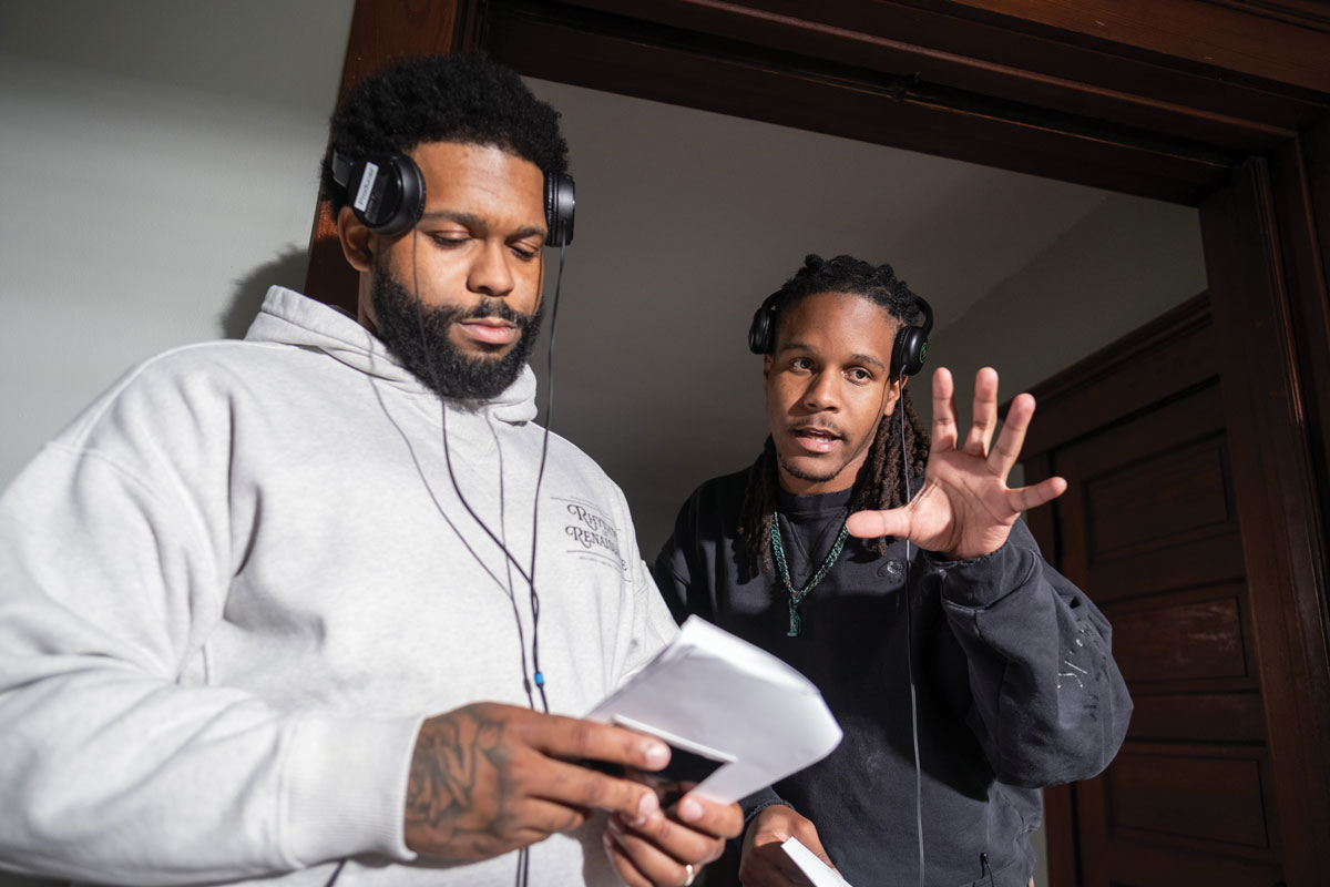 Julien Turner wears headphones pushed above his ears while Julien gestures and talks to him, also wearing his headphones pushed above his ears. They both wear sweatshirts and look focused.