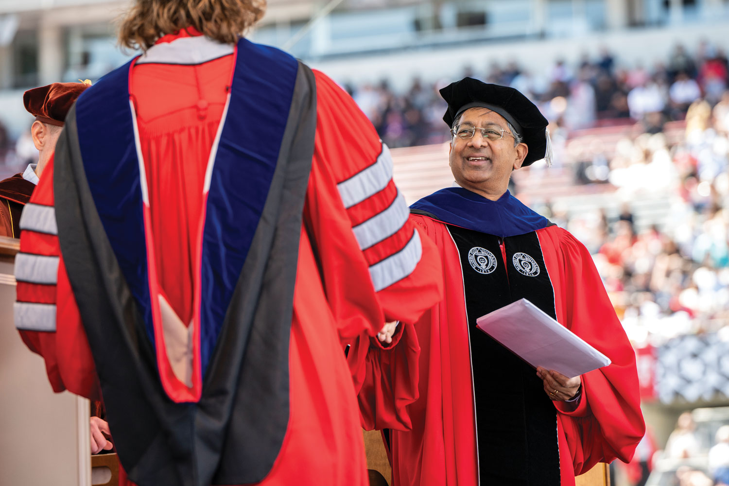 Dressed for commencement in ceremonial hat and robes, Bellamkonda smiles as he shakes the hand of a graduating student.