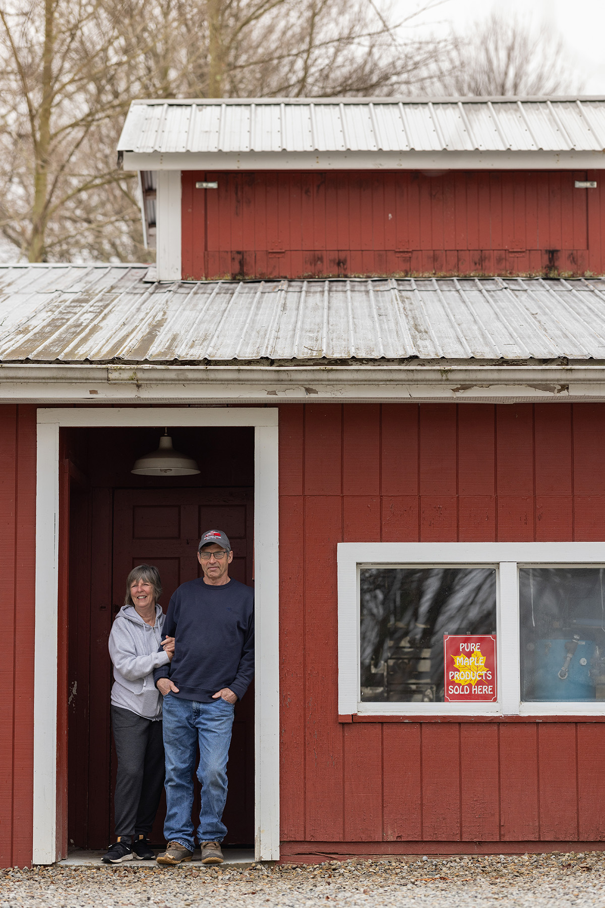 A husband and wife smile as they pose for a portrait in the doorway of of a building they use to cook maple syrup on their property. The building has a classic fall red-and-white design and is picturesque.