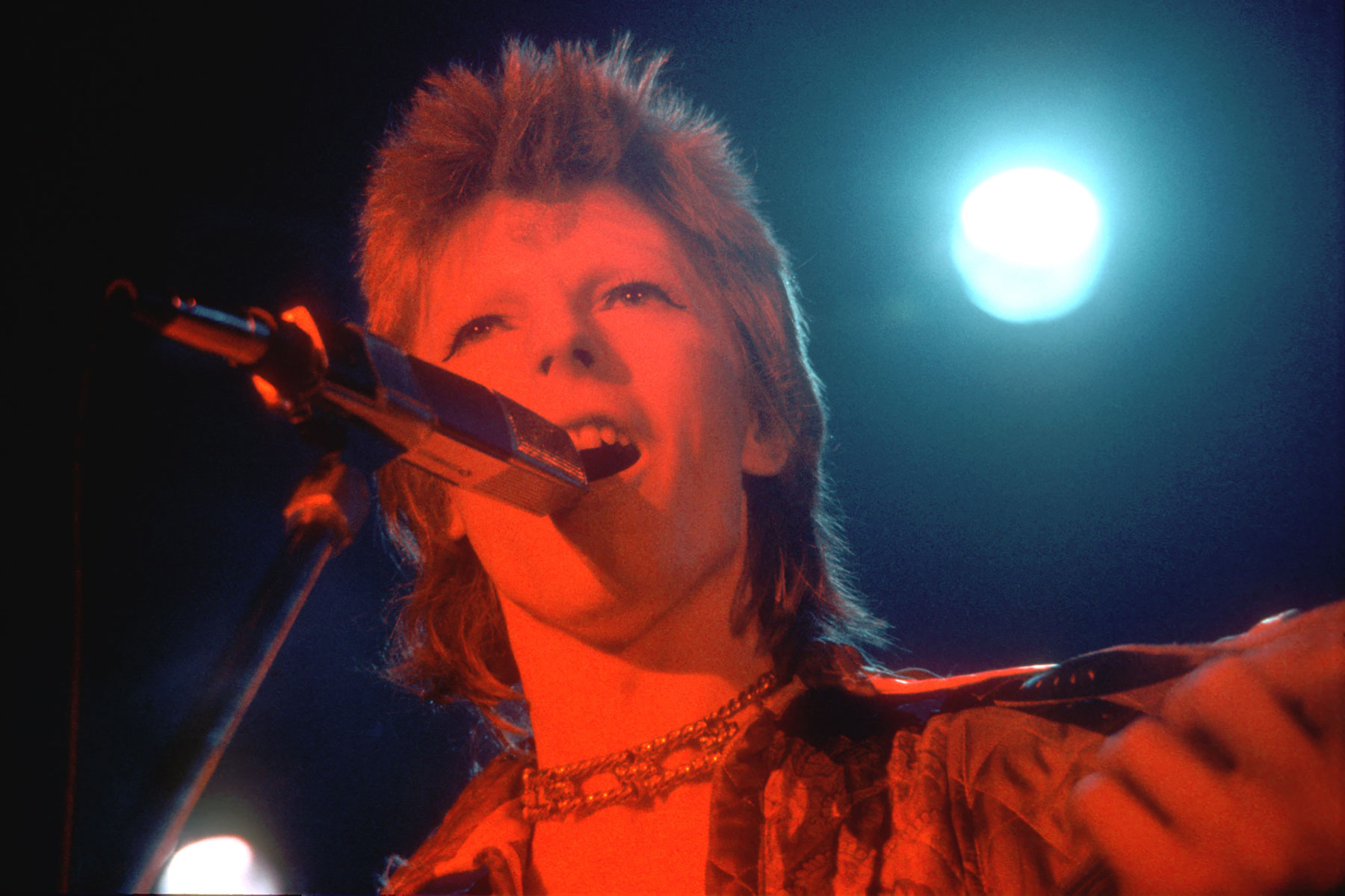 Red light shines on the face of an androgynous-appearing musician who sings into a mic.