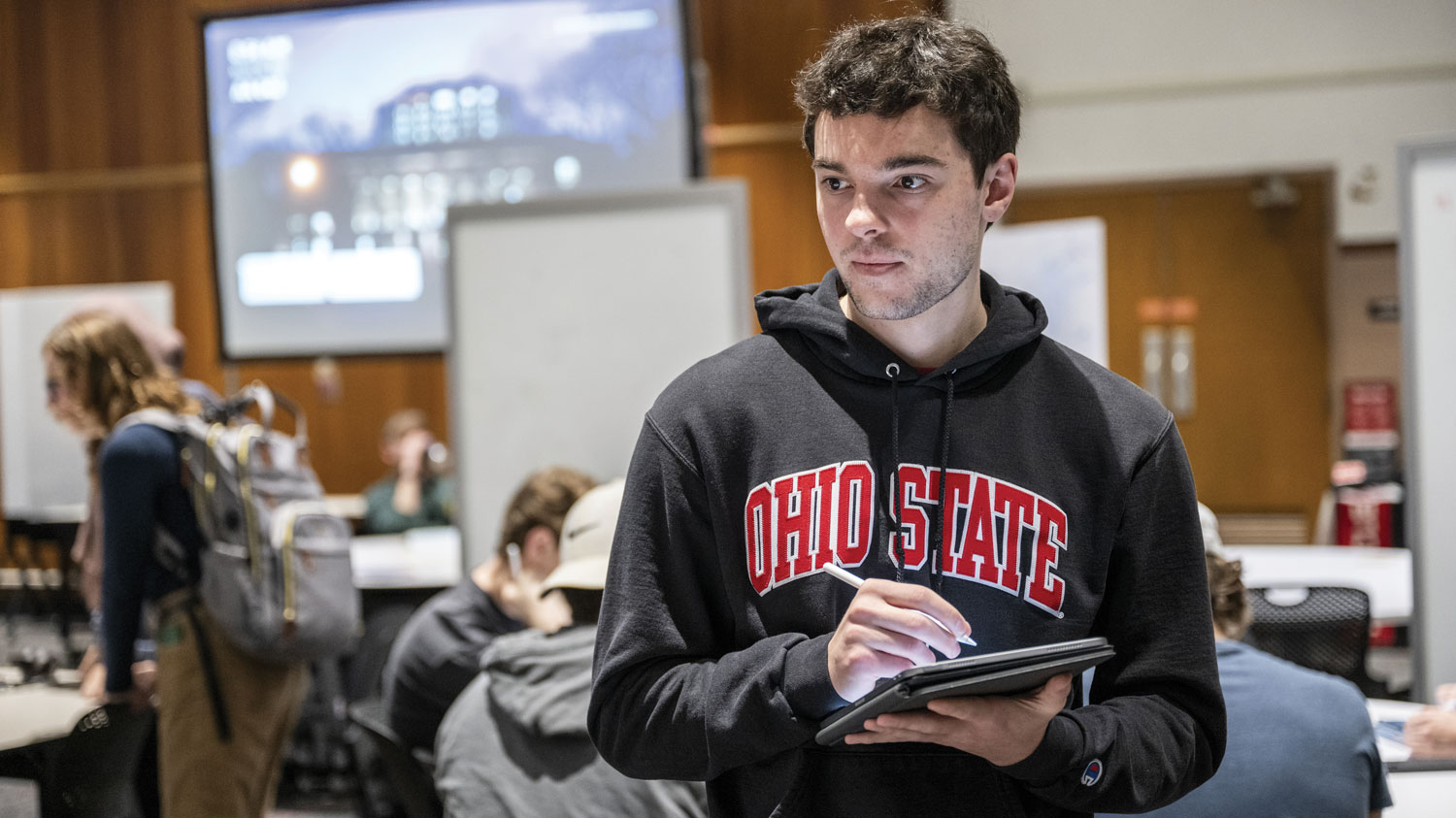 A student takes notes as classmates talk about his work. He wears an Ohio State sweatshirt and an intensely focused expression