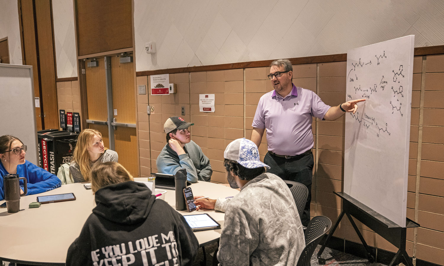 The chemistry teacher points to something on a white board as he talks with students at a small table. It’s a much more personalized experience than your usual lecture class.