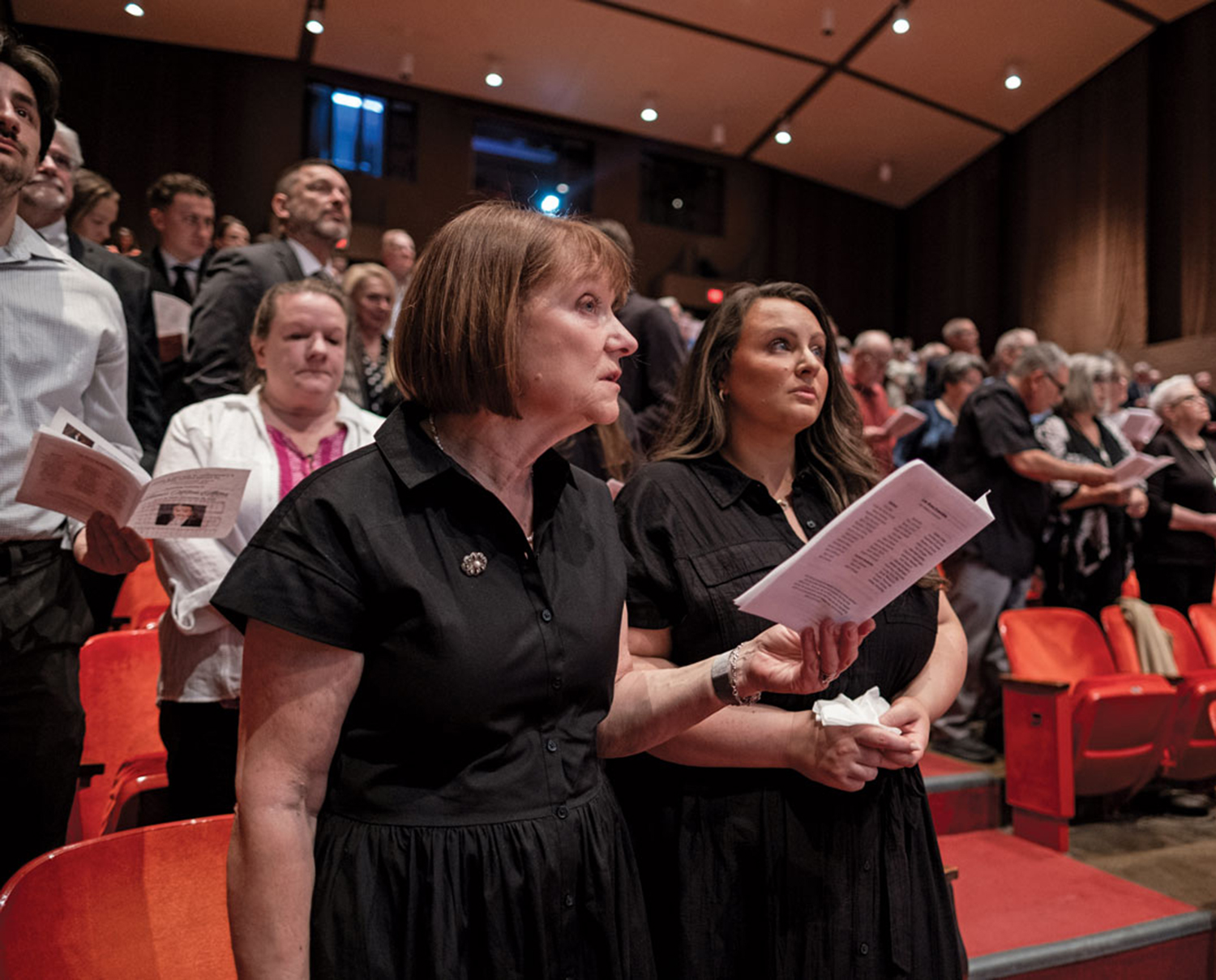 People stand in an auditorium during Akins’ celebration of life, holding paper programs and watching musicians perform on stage. The most prominent people in the photo are Akins’ widow and one of his daughters. They appear solemn.