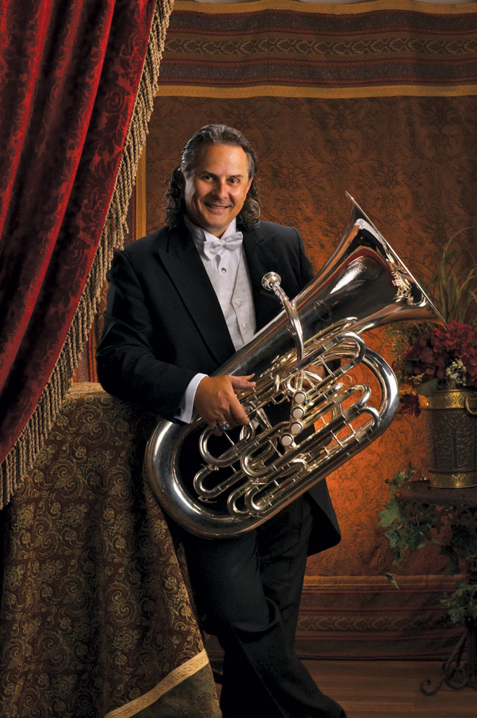 In his younger days, a long-haired Jim Akins smiles as he poses for an official portrait wearing a tux and holding his shiny tuba. A fancy background includes a velvet drape and brass pot of flowers.