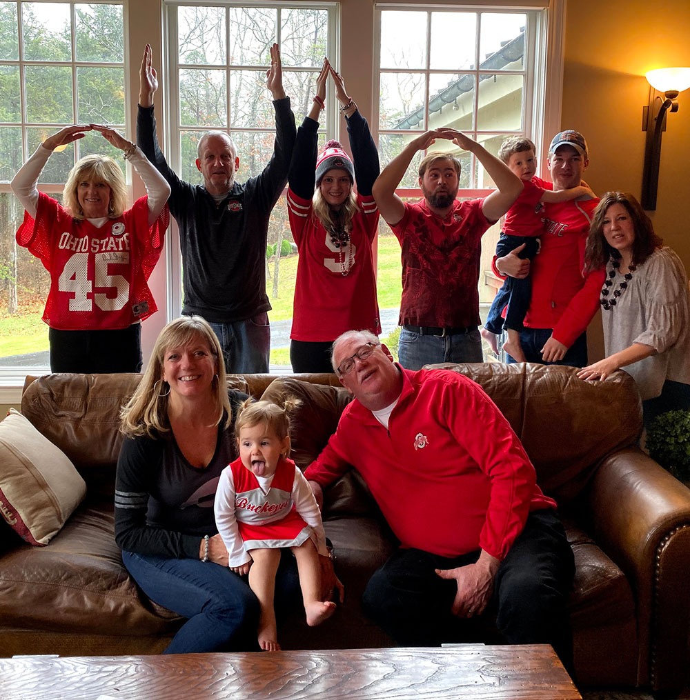 A family wearing Buckeyes jerseys or colors poses together in a living room, the back row standing behind a couch and making the O-H-I-O. The couple sitting on the couch hold a toddler in a Buckeyes cheerleader outfit. Everyone looks like their having fun.