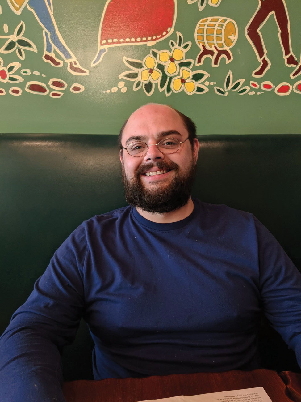 Eric Bretschneider smiles as he sits in a booth wearing a t-shirt and eyeglasses. He has dark hair, a beard and seems to be enjoying himself.