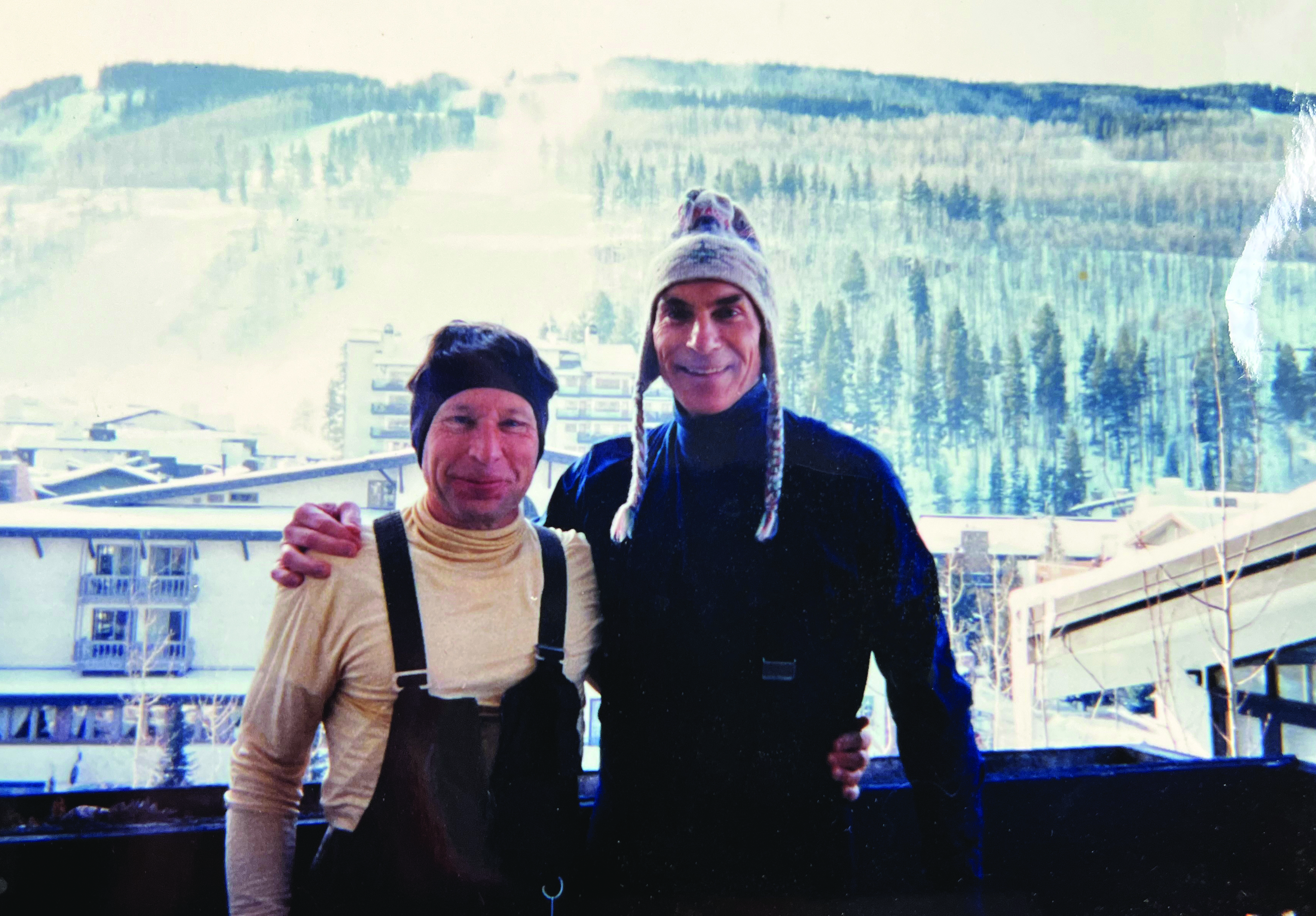 Two men pose in front on a wooded mountain while dressed in the layer of ski gear that goes under the coat. They're both smiling like they're amused.