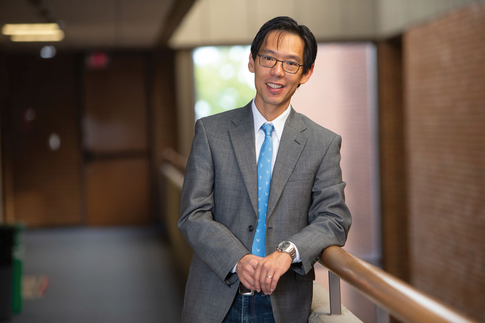 A man of Asian descent smiles as poses for this photo wearing a suit. He casually leans on a railing inside a university building. He looks like the kind of professor you'd want to take a class from--good natured and intelligent.