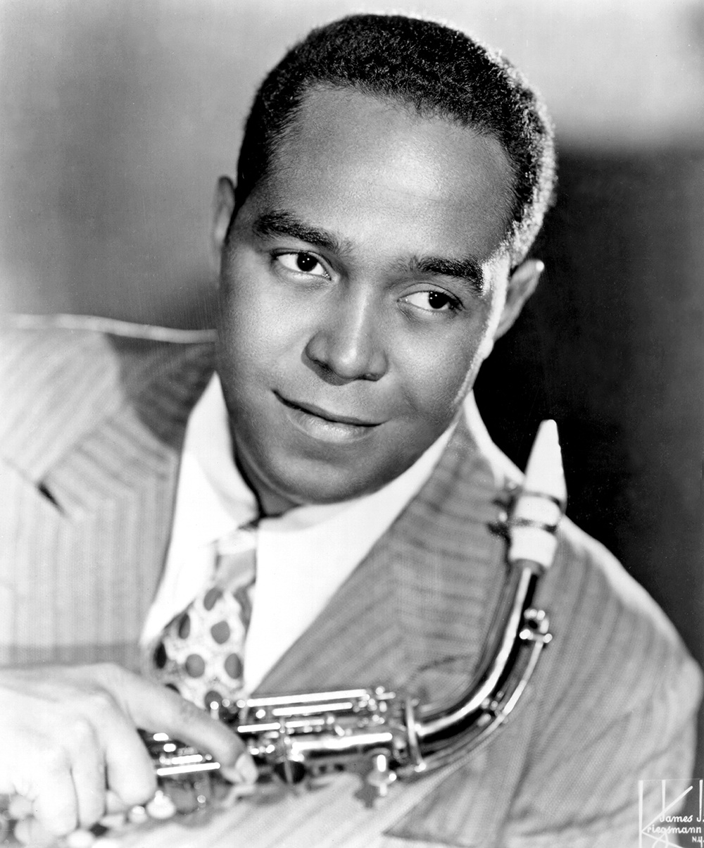 Name coming, a black man with neatly trimmed short hair, poses for a photo holding his saxophone. He wears a suit and looks very distinguished.  short