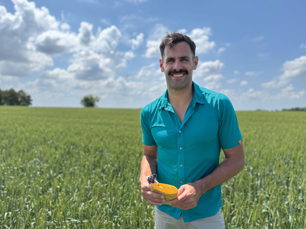 Jonathan Jacobs stands in a farm field on a beautiful day. He wears a short sleeved button-up shirt and holds sunglasses with rims the same color.