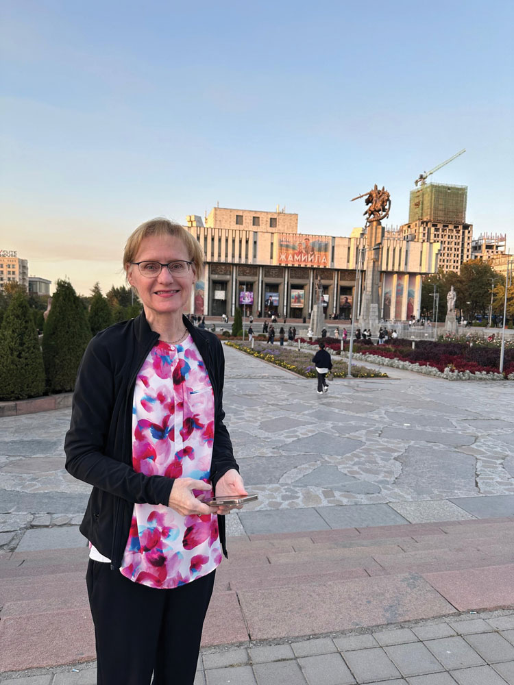 A snapshot shows Joni Tornwall in Kyrgyzstan, smiling as she stands in a plaza with other people. There's a low building in the background that seems to be a museum or governmental building. A banner with Cyrillic writing and illustrated people hangs from the roof. 