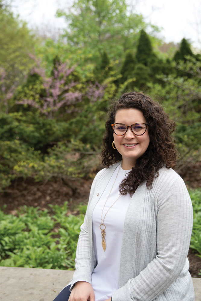 Mary Rodriguez sits for a portrait in an outdoor garden in a T-shirt and cardigan. She's a white woman genuinely smiling--you can tell from her crinkled eyes, behind her tortoise-shell-frame glasses.