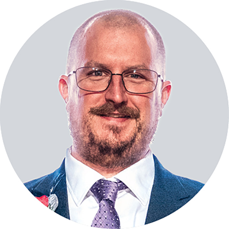 After getting the award, Andy May smiles for the camera. He wears glasses and a suit and has close-cropped hair and goatee.