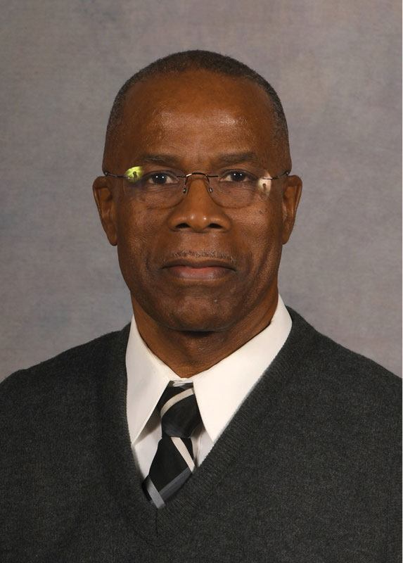 Melvin Pascal smiles with closed lips as he poses for an official headshot. He's a Black man wearing wire rimmed glasses and a v-necked sweater, tie and white button up.