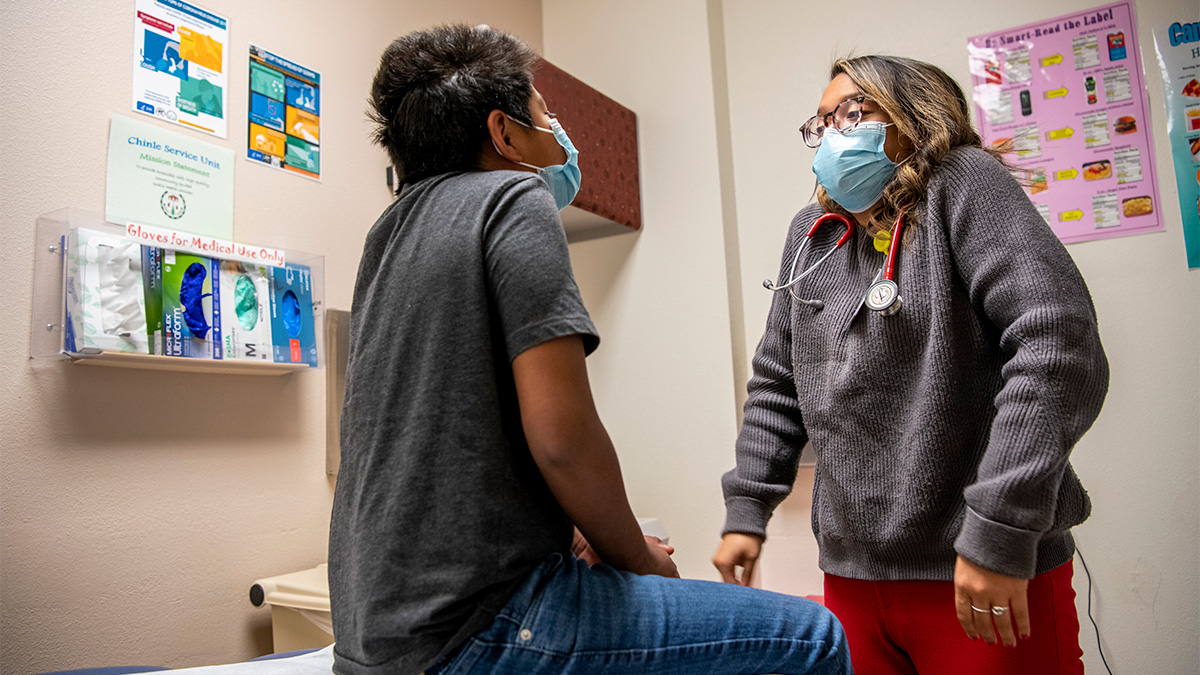 A young doctor in a surgical mask prompts a child patient to raise his shoulders during a medical exam