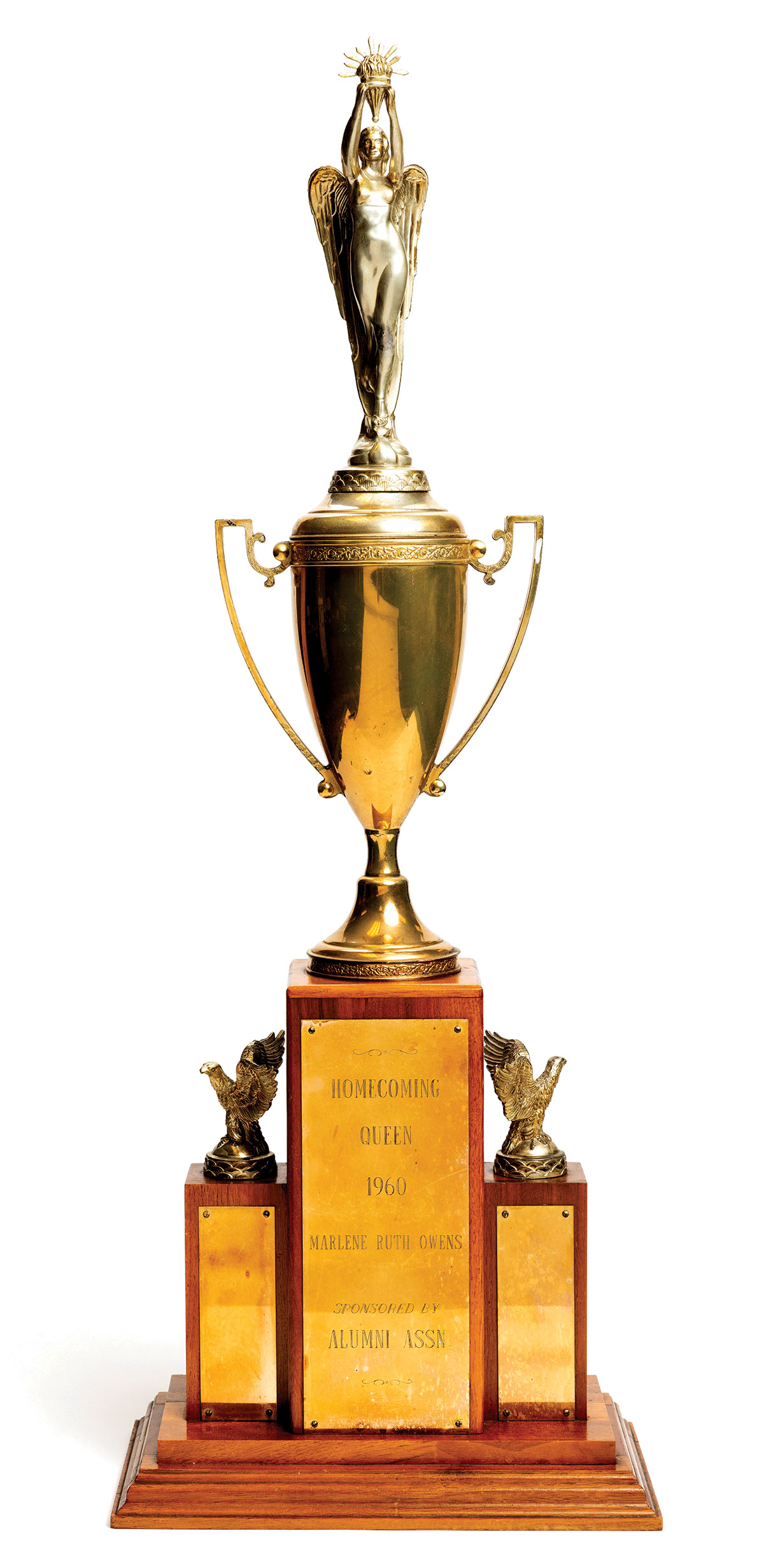 This is a large trophy made of polished wood and gold-colored metal. On the center, highest block, an angel holding what might be a torch stands atop a victory cup. On the shorter sides, eagles are perched. The central engraved plaque reads: Homecoming queen 1960, Marlene Ruth Owens.