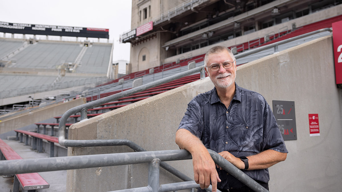 A white bearded man looks directly into the camera, like he’s about to  laugh along with a friend, in an aisle between ’Shoe stands.