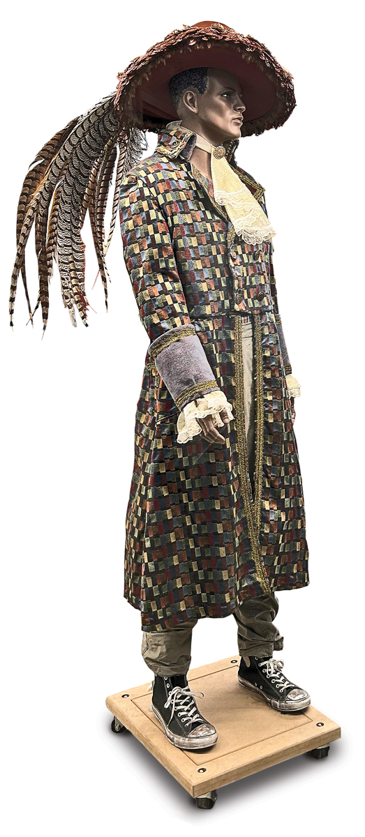 A gray mannequin wears a big hat with profuse feather cluster trailing to its waist; an old-fashioned, geometric-patterned coat that hangs to mid-shin; a lacy jabot; and jeans and old converse. The effect is a college student dressed for a play, perhaps something from Shakespeare.