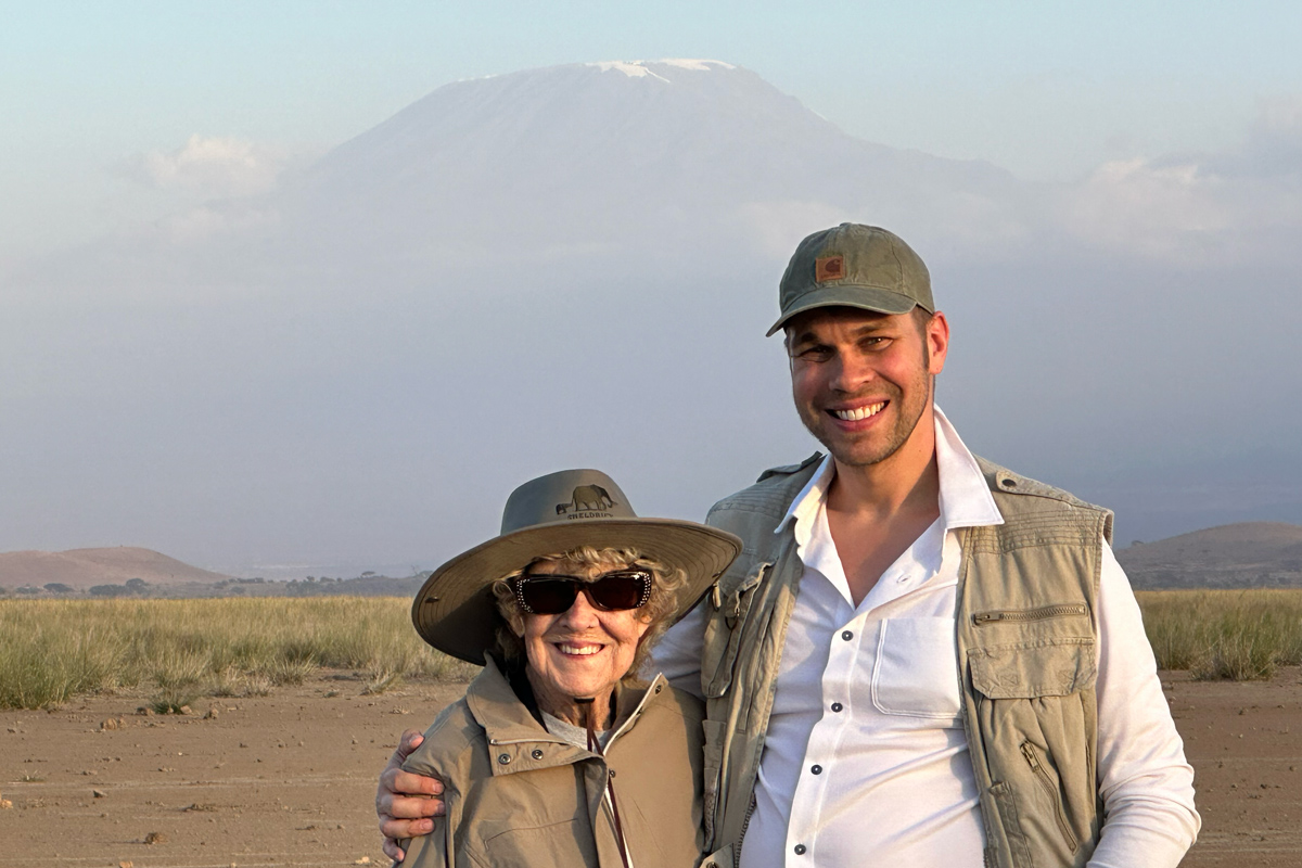 As Mount Kilimanjaro rises in the distance behind them, a white man stands with his arm wrapped around his much-shorter grandma. They wear khaki travelers’ clothes, hats and big smiles.