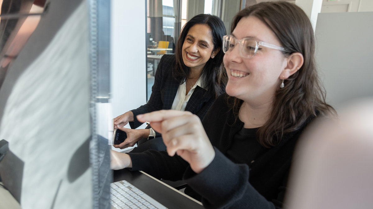 As Zoe Drellishak, a young woman wearing glasses, works at her computer, Bhakti sits beside her and laughs. Zoe points at the screen as she laughs as well. They’re in an office.
