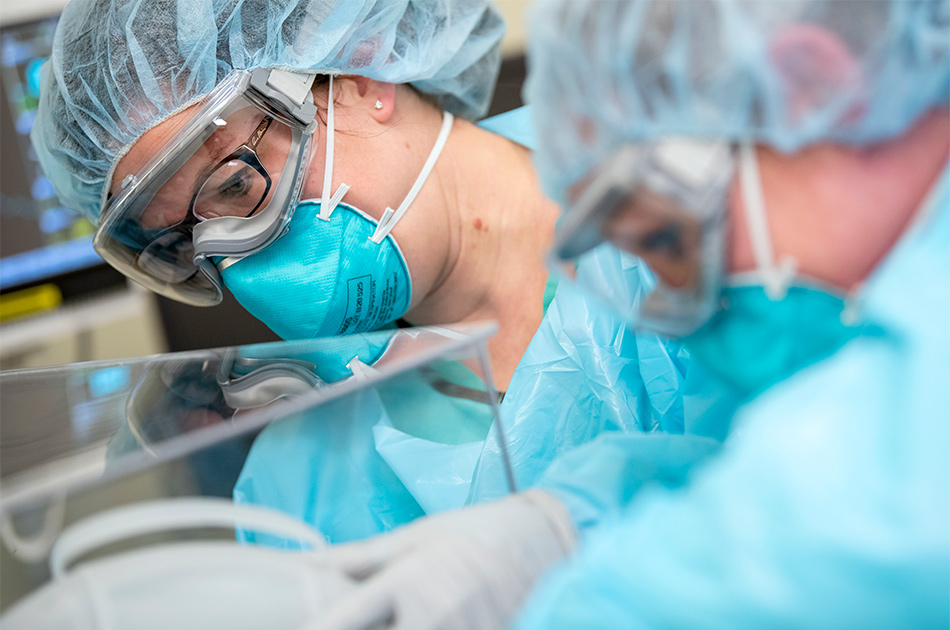 Closeup of two female nurses wearing hair nets, face masks, safety goggles, hospital gowns, and rubber gloves are touching a clear box