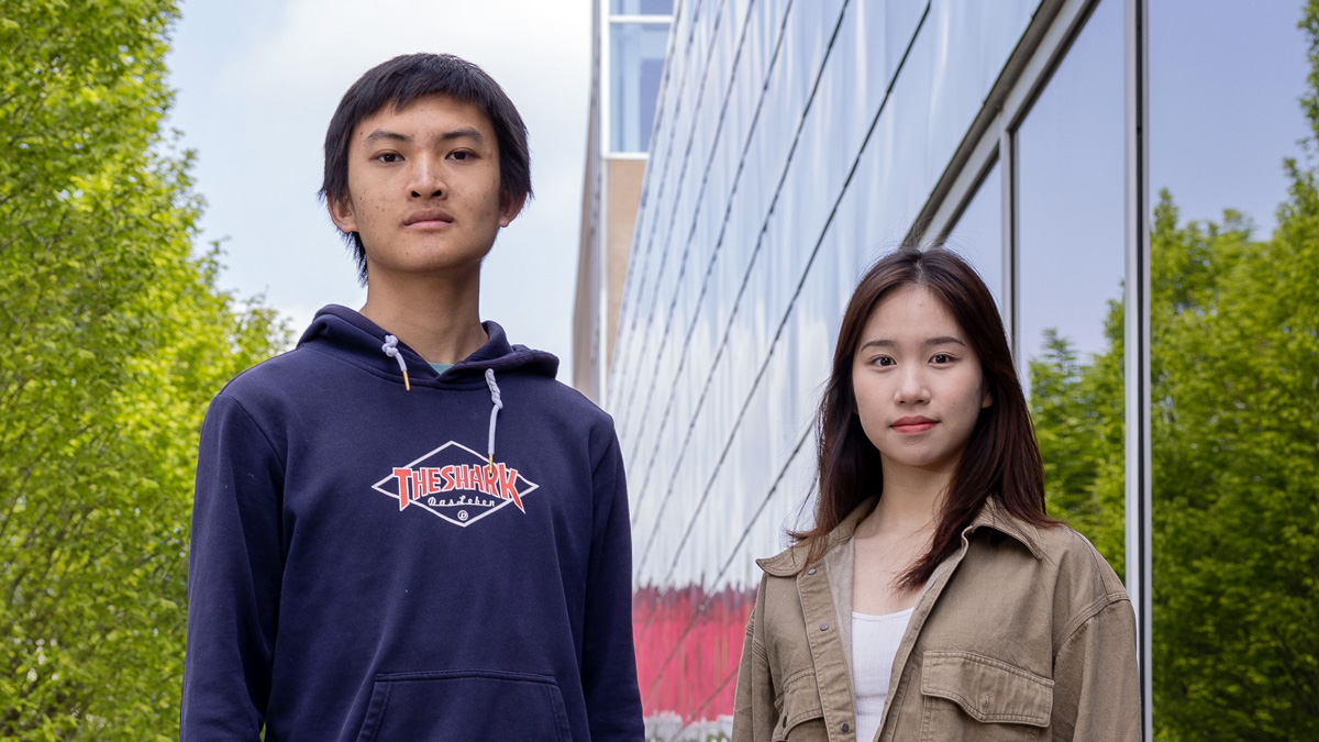 Two college students of Chinese descent pose for a photo next to Ohio State’s recreation building. A young man on the left wears a sweatshirt and sweatpants and a serious look. The student on the right is a young woman with long hair and a slight smile.
