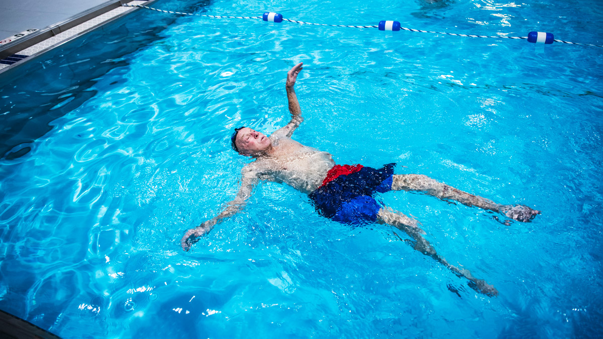 Merle Lashey Jr., wearing swim trunks and a cap, floats on his back in an indoor pool. On the other side of a rope divider, women are in a line, as if taking an exercise class.