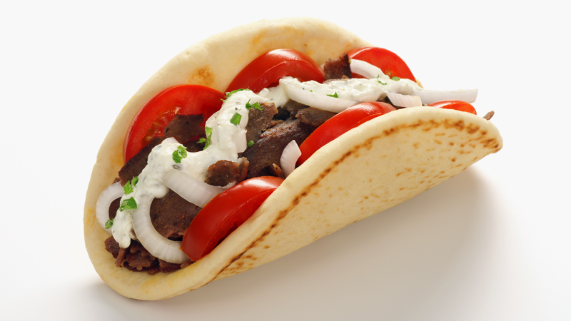 A close-up photo of a greek gyro — sliced lamb, tomato, tzatziki sauce, lettuce, on a folded pita bread