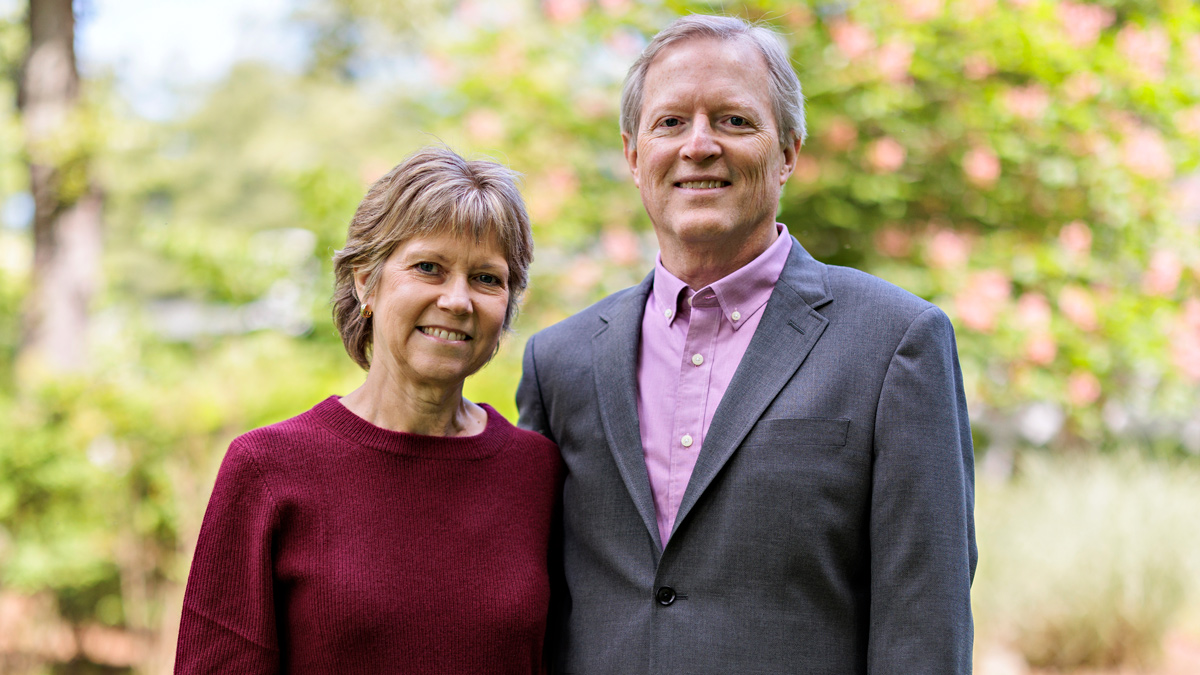 A middle-aged couple stand closely while holding hands. The woman smiles big enough that you can see her teeth; her husband has a closed-lip smile, but both have smiling eyes, too. They’re outside on a pretty day.