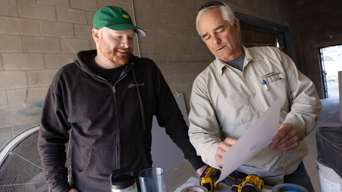 Standing over a hip-height table in a big room with concrete-clock walls, two white men look at a piece of paper one is holding to point something out. The customer is Adam Benner, and he is smiling.
