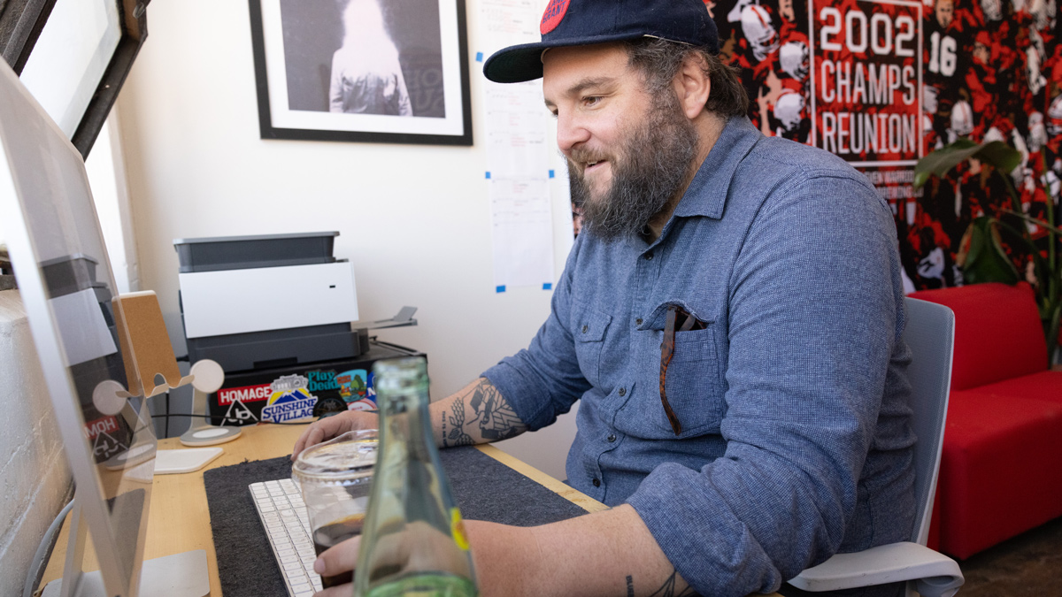 Walt Keys, a bearded white man, sits at a computer holding an iced coffee in one hand and his computer mouse in the other. He’s smiling over something on the screen that can’t be seen. On the wall behind him is a giant Ohio State football poster, a shot of the players and fans on the field, and text says 2002 champs reunion.