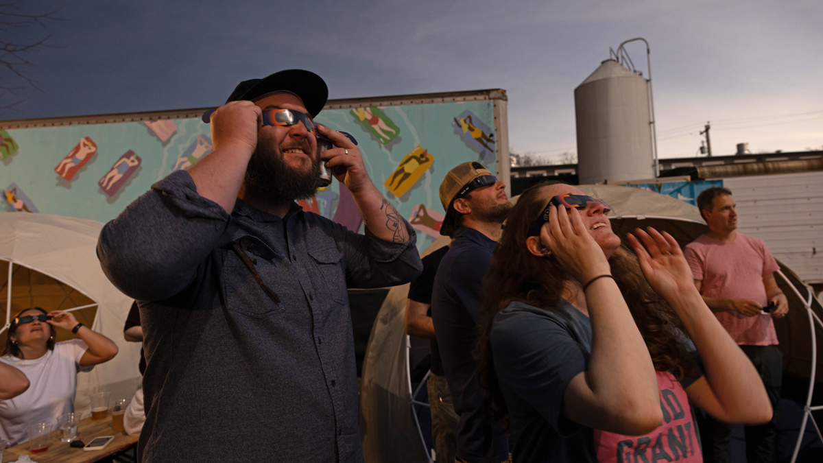 Walt and others wearing eclipse glasses marvel as they look up at the darkened sky, watching the moon cover the sun. The light in this photo shows how the eclipse made it much darker during daytime.