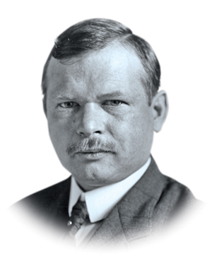 A white man with neatly parted hair and a faint mustache looks just off camera. He wears a suit and an expression that says he’s considering something in this black and white portrait.
