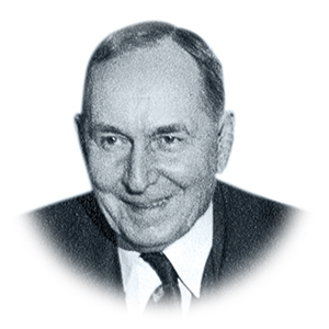 An older, balding man smiles as if he is delighted by something. He’s wearing a suit but his tie is slightly crooked in this black and white photo.
