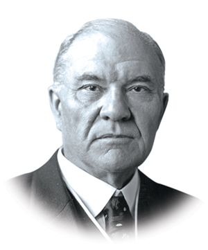 A white man who is starting to bald stares just off camera. He wears a suit and a serious expression in this black and white portrait.