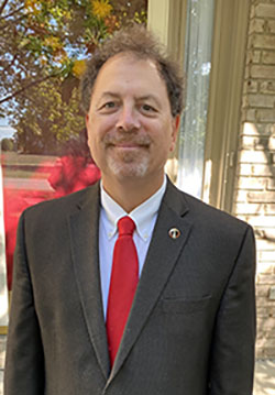 Retired Lt. Col. Richard Hughes poses for a photo wearing a neat civilian suit and a smile. He has friendly eyes and curly hair.