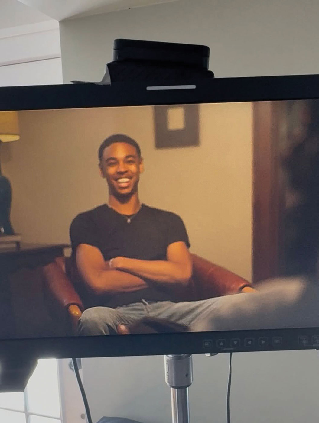 Reign Winston sits in a chair and smiles with arms crossed, as seen through a monitor on a production set.