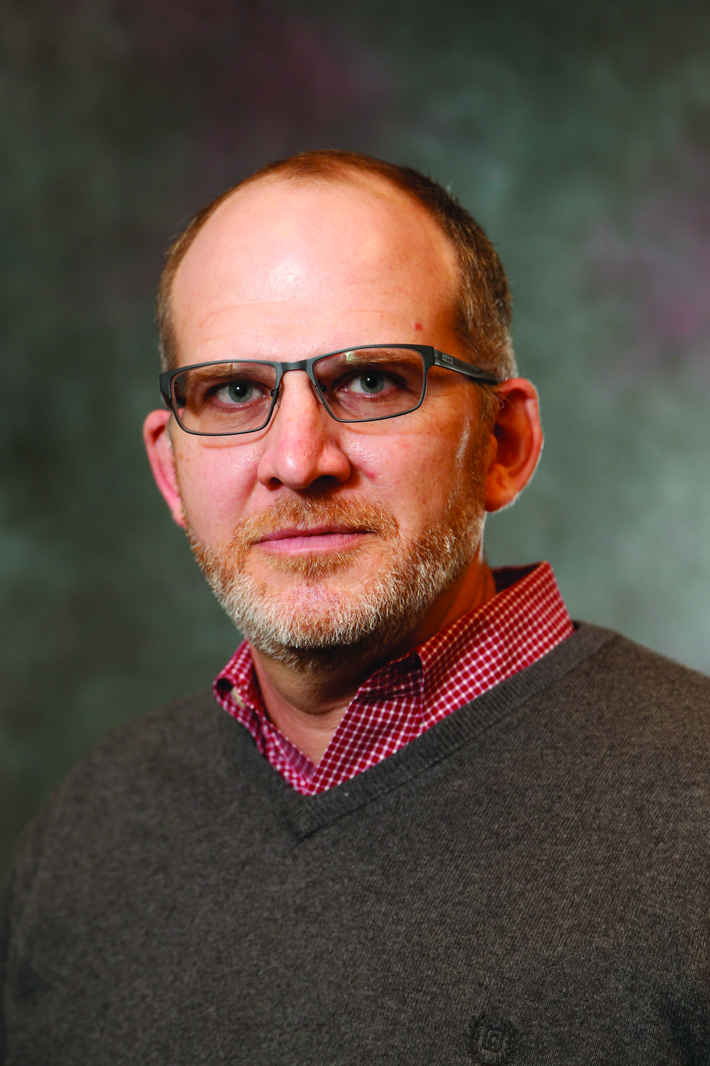 Zac Schultz poses for an official portrait. He's a white man wearing glasses, a v-neck sweater and a checked shirt.