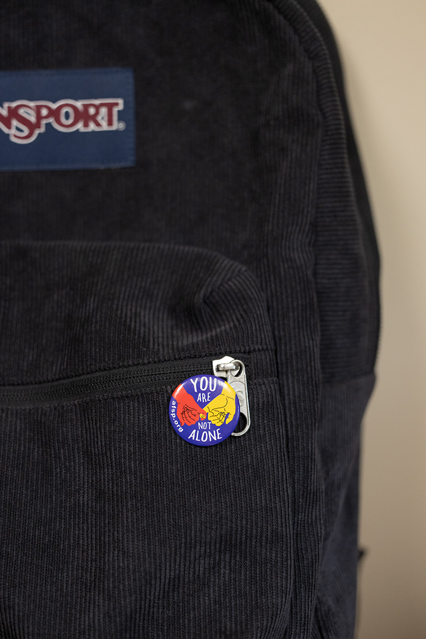 On a black, corduroy backpack, a colorful pin shows two hands with interlinked pinkies. "You are not alone," it says, along with afsp.org, the URL for the American Foundation for Suicide Prevention.