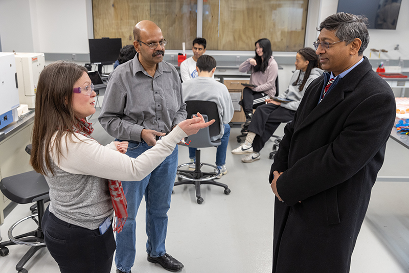 In an education lab, Ravi Bellamkonda intently watches a white woman speaking to him. She has long hair and wears safety goggles. Standing with them is another teacher, an older man of Indian descent.