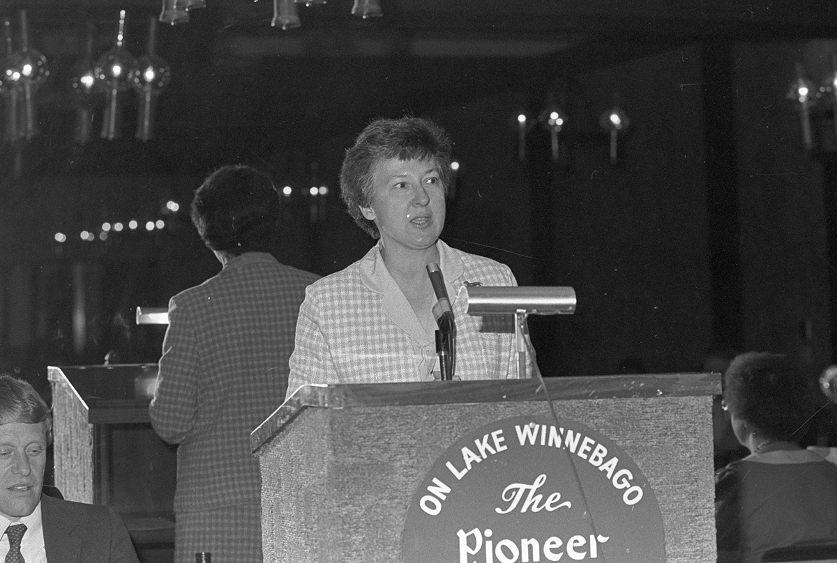 Helen Briwa speaks at a podium in an old black and white photo. She wears a checked jacket and a mirror behind her reflects her back and, faintly, the audience.