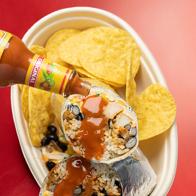 A burrito that has been cut in half is shown with open-ends up. Hot sauce is being poured across the visible rice, chorizo, black beans and potatoes. On the plate next to the burrito are corn chips.
